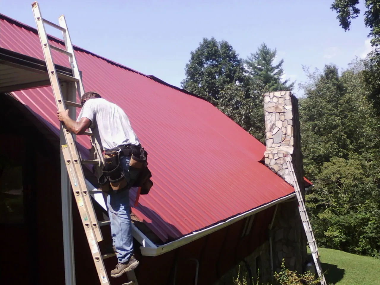 Roofer on a ladder beside a red metal roof and stone chimney — Soffit & Fascia Repair by Roofing Brothers in Woodberry Forest